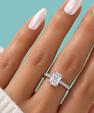 A close-up of a hand wearing a silver engagement ring with a cushion-cut diamond and a pavé band, set against a solid teal background.