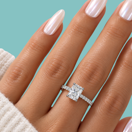 A close-up of a hand wearing a silver engagement ring with a cushion-cut diamond and a pavé band, set against a solid teal background.