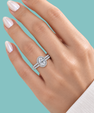 A close-up of a hand wearing a silver halo ring with a round-cut center stone and pavé band, set against a solid light teal background.