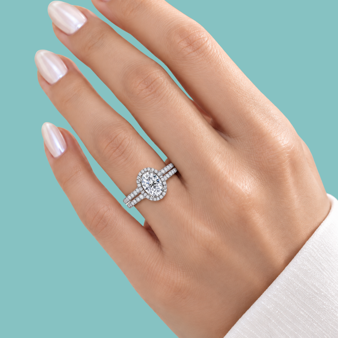A close-up of a hand wearing a silver halo ring with a round-cut center stone and pavé band, set against a solid light teal background.