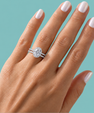 A close-up of a hand with pale pink manicured nails wearing a silver-toned double-band ring with a large oval-cut center stone and smaller stones along the band, set against a solid teal background.