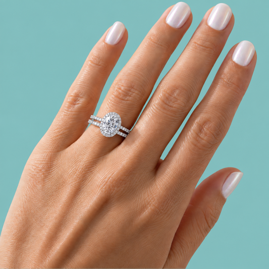 A close-up of a hand with pale pink manicured nails wearing a silver-toned double-band ring with a large oval-cut center stone and smaller stones along the band, set against a solid teal background.