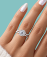 A close-up of a hand wearing a silver halo engagement ring with a round-cut center stone and matching band, set against a solid teal background.