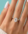 A close-up of a hand wearing a yellow gold ring with an oval-cut clear center stone and small side stones, set against a solid turquoise background.