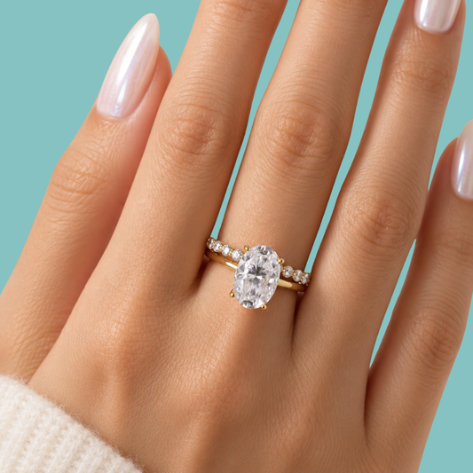 A close-up of a hand wearing a yellow gold ring with an oval-cut clear center stone and small side stones, set against a solid turquoise background.