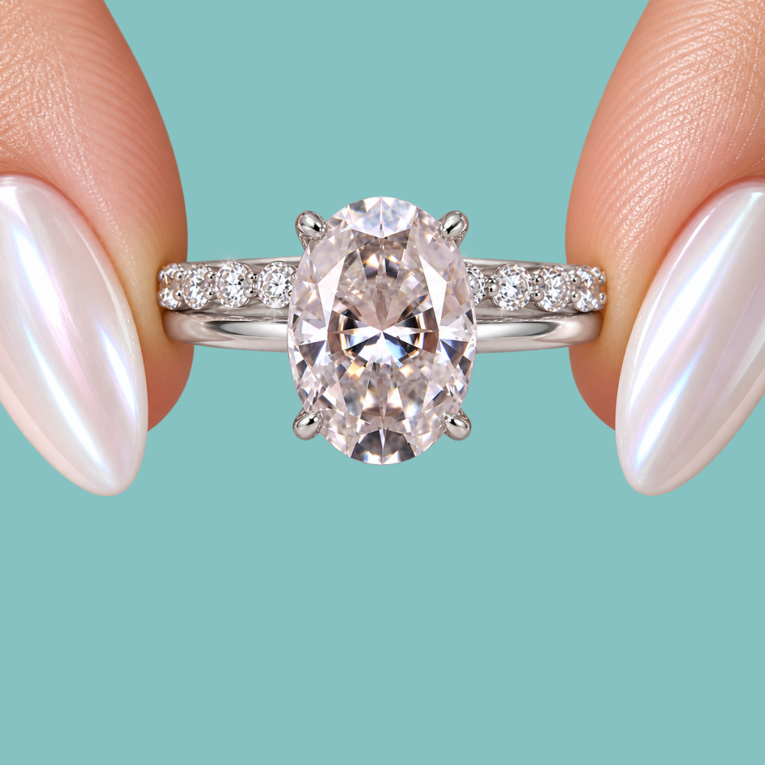An oval-cut diamond engagement ring with a silver-toned band accented by small side diamonds, held between two fingers with iridescent nail polish against a solid teal background.