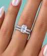 A close-up of a hand wearing a silver engagement ring with a rectangular clear center stone and matching pavé bands, set against a solid teal background.