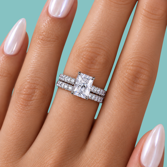 A close-up of a hand wearing a silver engagement ring with a rectangular clear center stone and matching pavé bands, set against a solid teal background.