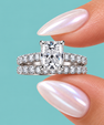 A radiant cut diamond engagement ring with a matching pavé band, held between fingers with iridescent nail polish against a solid turquoise background.