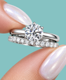 A close-up of a silver engagement ring with a round brilliant-cut diamond and a matching diamond band, held between fingers against a solid teal background.