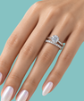 A close-up of a hand wearing a silver engagement ring with a round-cut center stone and a matching diamond band, set against a solid teal background.