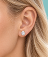 A close-up of a woman's ear wearing a small oval stud earring with a clear gemstone in a gold setting, against a solid teal background.