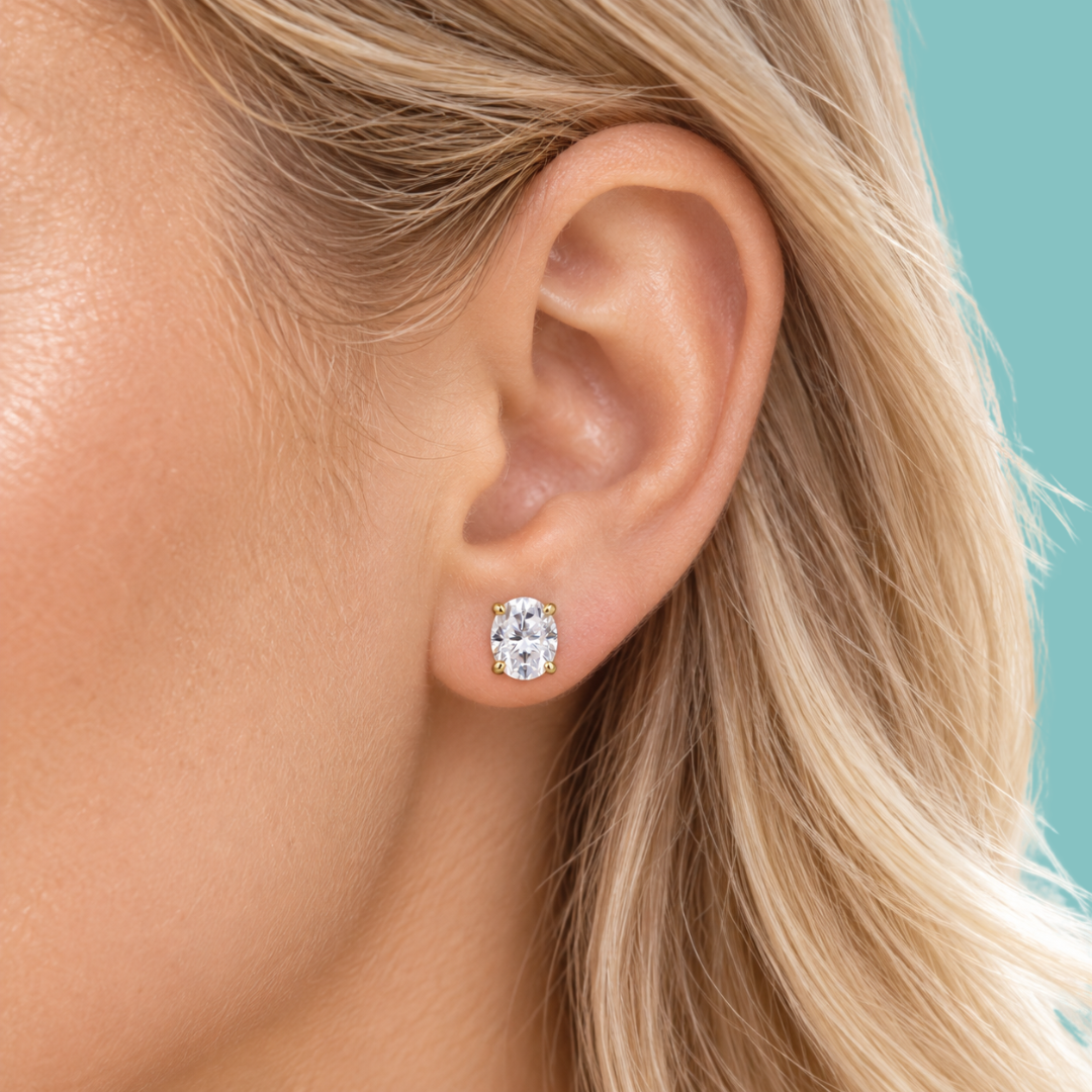 A close-up of a woman's ear wearing a small oval stud earring with a clear gemstone in a gold setting, against a solid teal background.