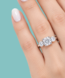 A close-up of a hand wearing a three-stone diamond engagement ring with a silver-colored band, set against a solid teal background.