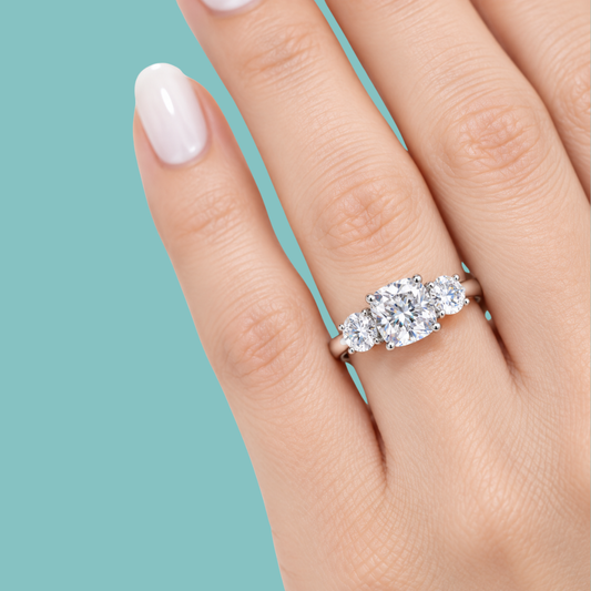 A close-up of a hand wearing a three-stone diamond engagement ring with a silver-colored band, set against a solid teal background.