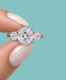 A three-stone diamond engagement ring held between fingers against a solid turquoise background. The ring features a large cushion-cut center diamond flanked by two round side diamonds, set in a silver-colored metal.