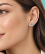 A close-up of a woman's ear wearing a small square cushion-cut diamond stud earring in a silver-toned setting, against a solid teal background.