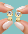 Gold stud earrings with moissanite clear gemstones, shown on fingertips with pale pink manicured nails against a solid light blue background.