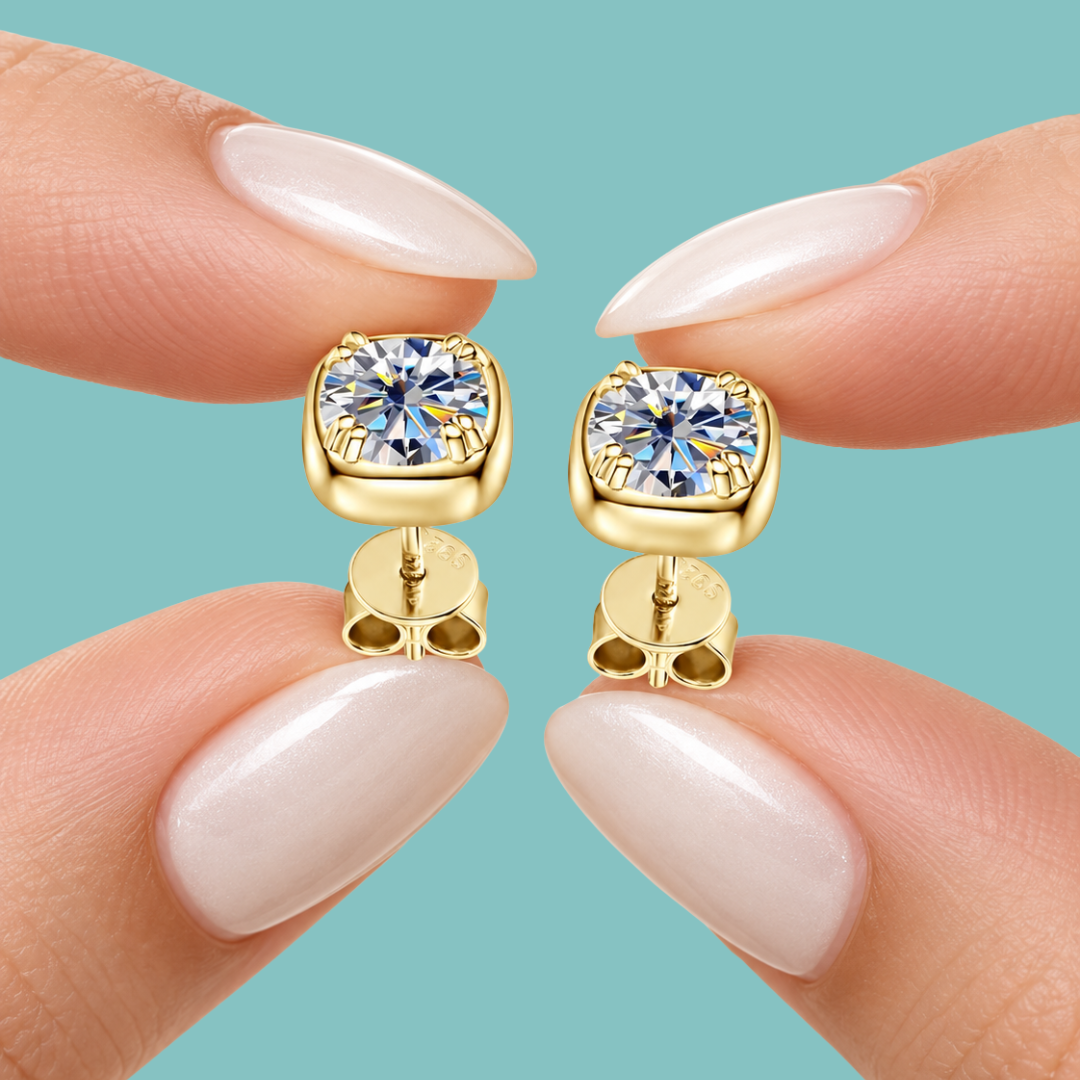 Gold stud earrings with moissanite clear gemstones, shown on fingertips with pale pink manicured nails against a solid light blue background.