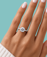 A close-up of a hand wearing a round-cut diamond engagement ring on a silver band, set against a solid teal background.