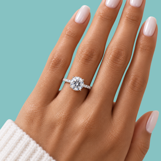 A close-up of a hand wearing a round-cut diamond engagement ring on a silver band, set against a solid teal background.