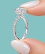 A close-up of a silver diamond ring held between two manicured fingers against a solid light teal background.