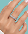 A close-up of a hand wearing a silver eternity ring set with clear round-cut gemstones, set against a solid teal background.