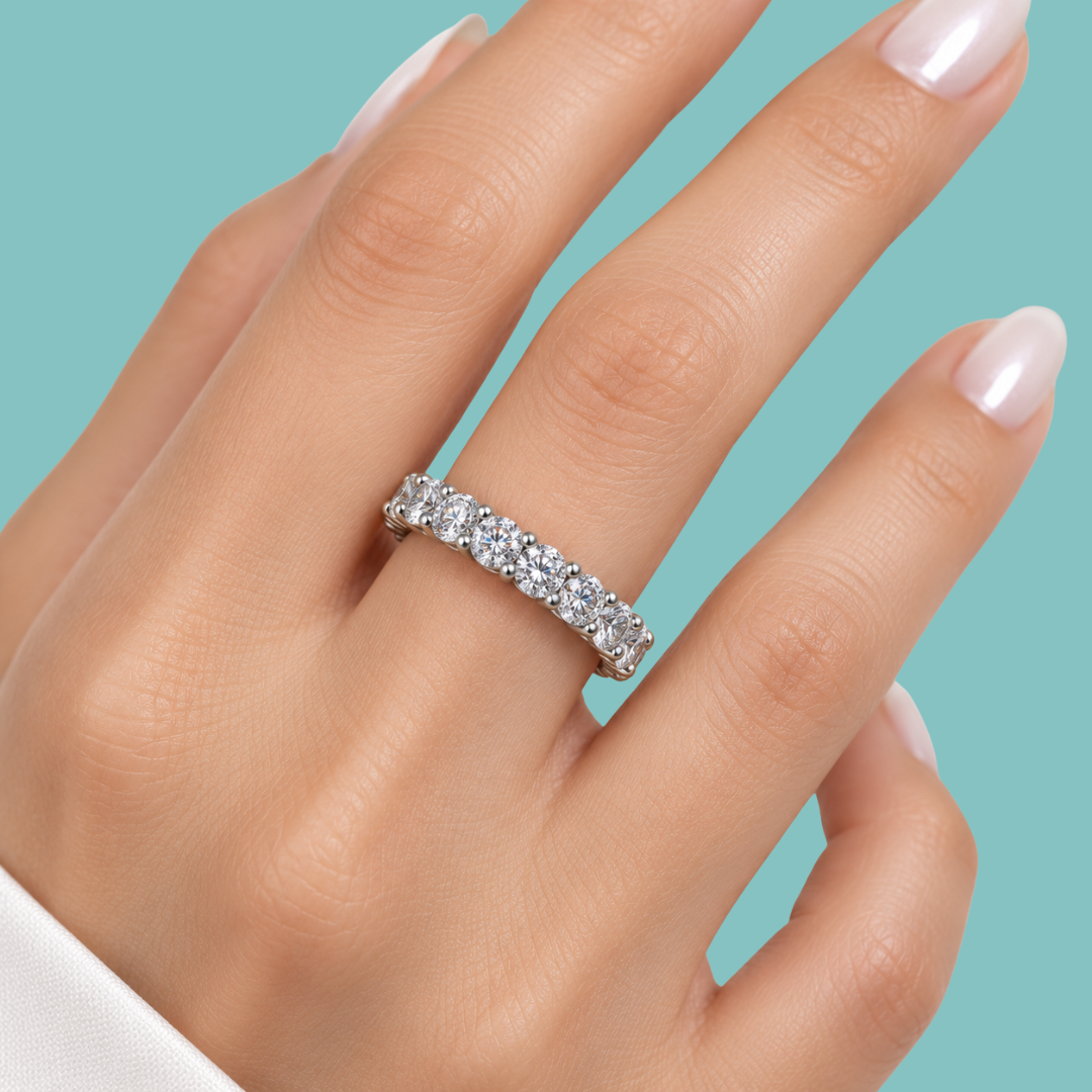 A close-up of a hand wearing a silver eternity ring set with clear round-cut gemstones, set against a solid teal background.