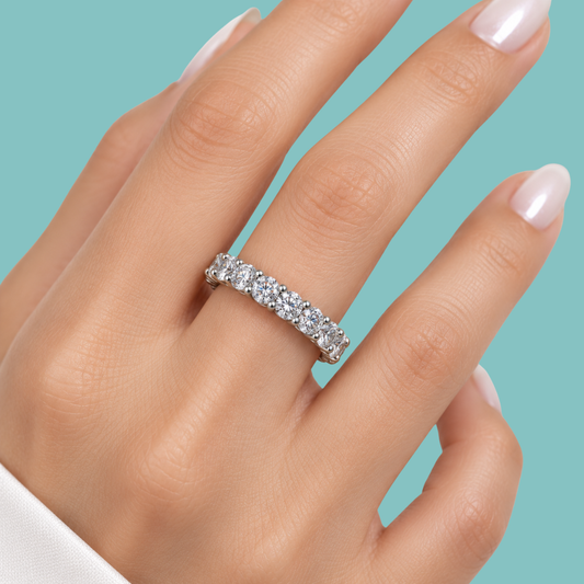 A close-up of a hand wearing a silver eternity ring set with clear round-cut gemstones, set against a solid teal background.
