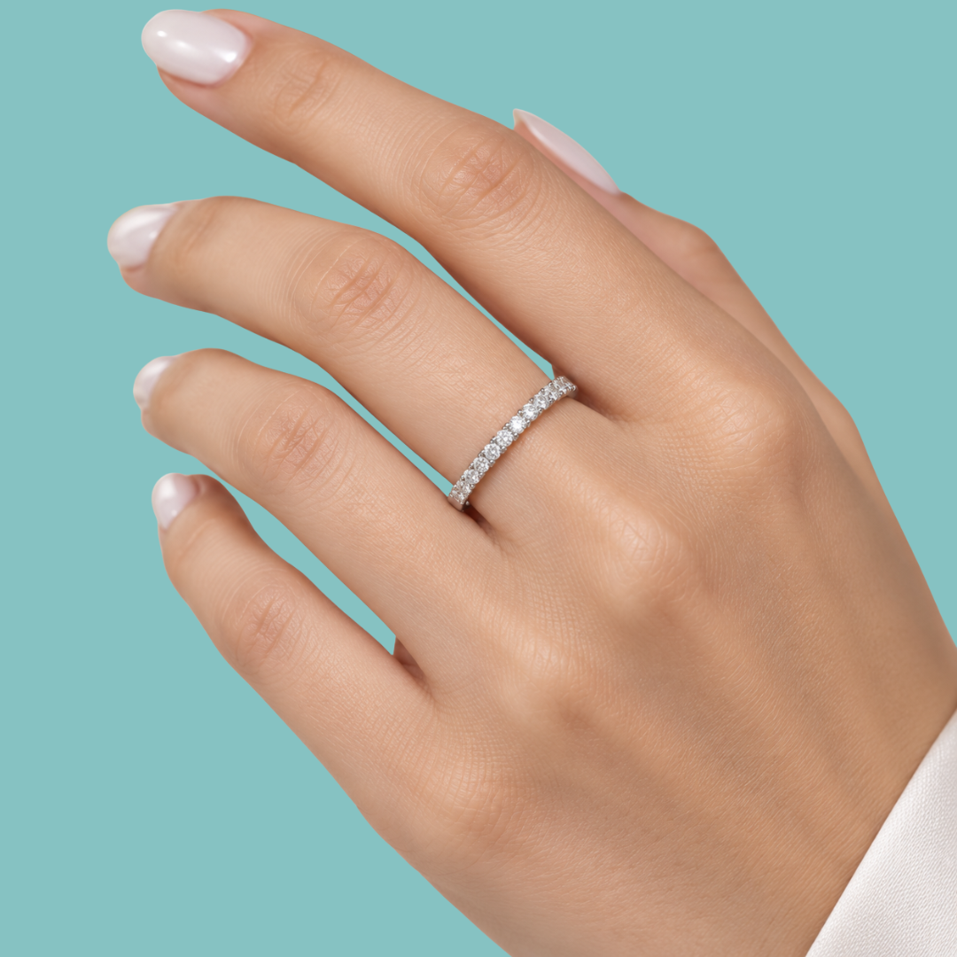 A close-up of a hand wearing a thin silver eternity ring set with small clear stones, against a solid light teal background.