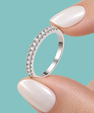 A close-up of a silver ring with a row of clear cubic zirconia stones, held between two fingers with pale pink manicured nails against a solid teal background.