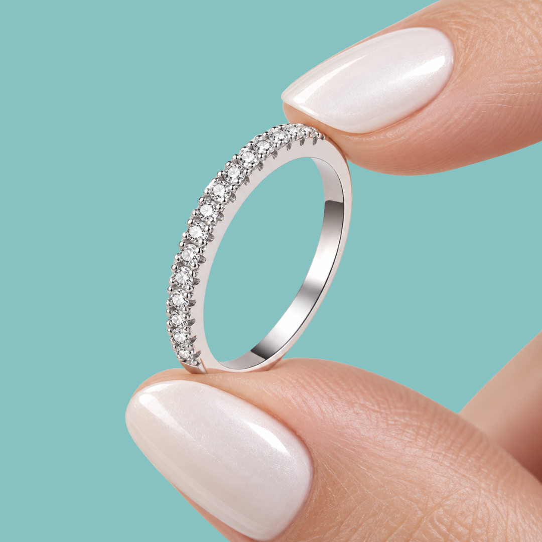 A close-up of a silver ring with a row of clear cubic zirconia stones, held between two fingers with pale pink manicured nails against a solid teal background.