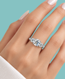 A close-up of a hand wearing a silver three-stone diamond engagement ring against a solid teal background.