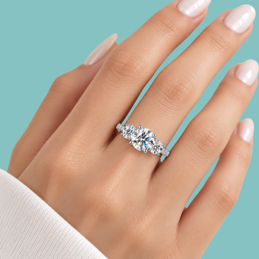 A close-up of a hand wearing a silver three-stone diamond engagement ring against a solid teal background.