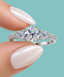 A silver three-stone diamond engagement ring held between fingers against a solid light teal background.