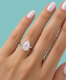 A pear-cut diamond ring on a silver band, shown worn on a hand with pale pink manicured nails against a solid light blue background.