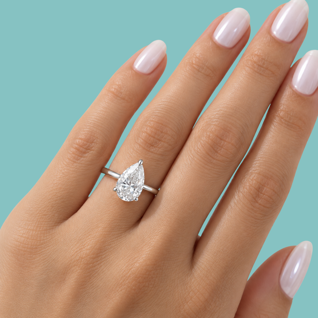 A pear-cut diamond ring on a silver band, shown worn on a hand with pale pink manicured nails against a solid light blue background.