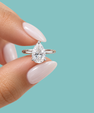 A close-up of a hand with pale pink manicured nails holding a silver ring with a pear-cut clear gemstone against a solid light blue background.