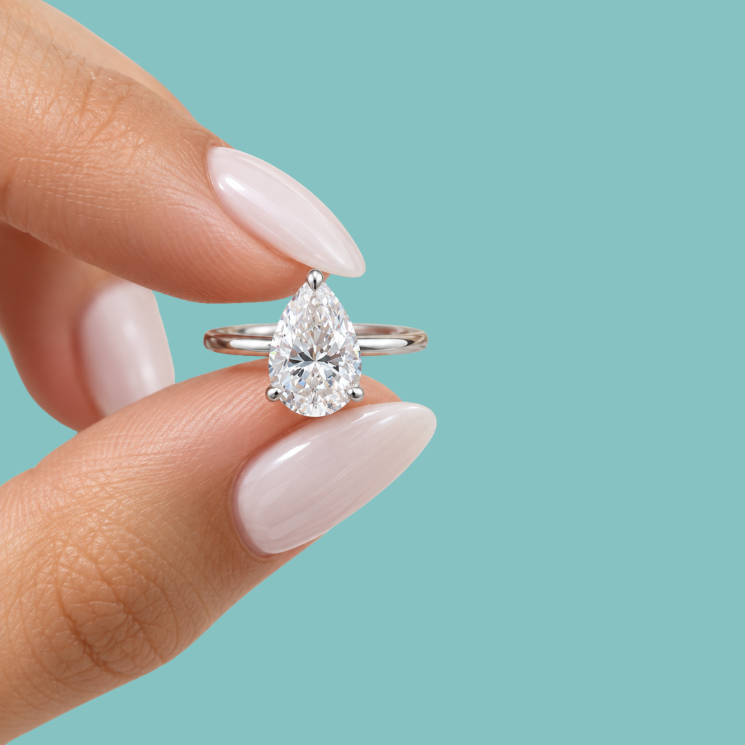 A close-up of a hand with pale pink manicured nails holding a silver ring with a pear-cut clear gemstone against a solid light blue background.