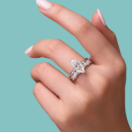 A close-up of a hand wearing a silver-toned ring with a marquise-cut center stone and smaller side stones, set against a solid turquoise background.