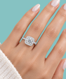 A close-up of a hand wearing a silver-toned engagement ring with a cushion-cut diamond center and a halo of smaller diamonds, set against a solid light blue background.