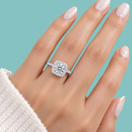 A close-up of a hand wearing a silver-toned engagement ring with a cushion-cut diamond center and a halo of smaller diamonds, set against a solid light blue background.
