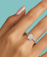 A close-up of a hand wearing a silver engagement ring with an emerald-cut diamond and a pavé band, set against a solid light blue background.