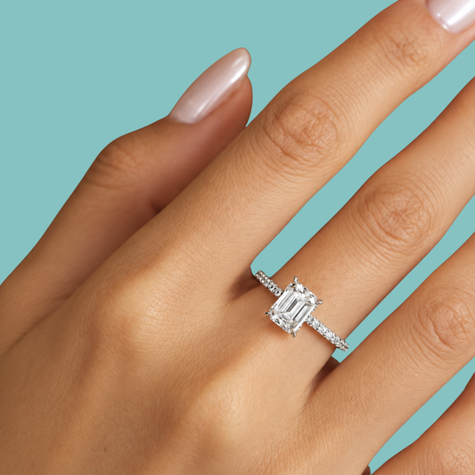 A close-up of a hand wearing a silver engagement ring with an emerald-cut diamond and a pavé band, set against a solid light blue background.