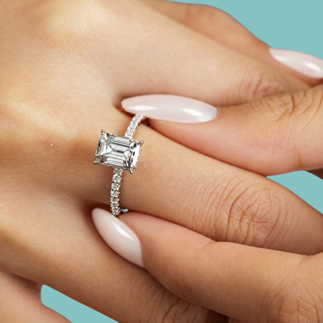 A close-up of a hand with pale pink manicured nails wearing a silver engagement ring with an emerald-cut clear center stone and a pavé band, set against a solid teal background.