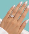 A close-up of a hand with long, pale pink manicured nails wearing a silver-toned ring with a round-cut clear gemstone, set against a solid turquoise background.