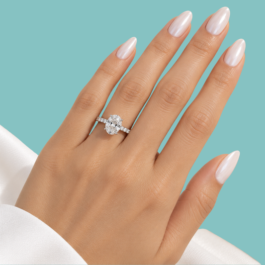 A close-up of a hand with long, pale pink manicured nails wearing a silver-toned ring with a round-cut clear gemstone, set against a solid turquoise background.