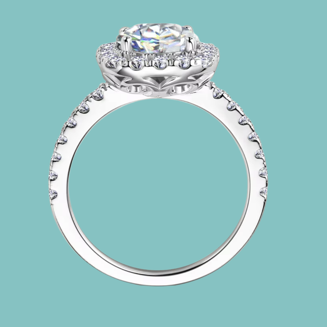 A silver-toned halo engagement ring with a round-cut center stone and small accent stones along the band, shown on a solid light teal background.