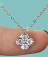 A silver-tone four-flower pendant necklace with four clear round-cut stones, shown resting on a person's fingertip against a solid turquoise background.