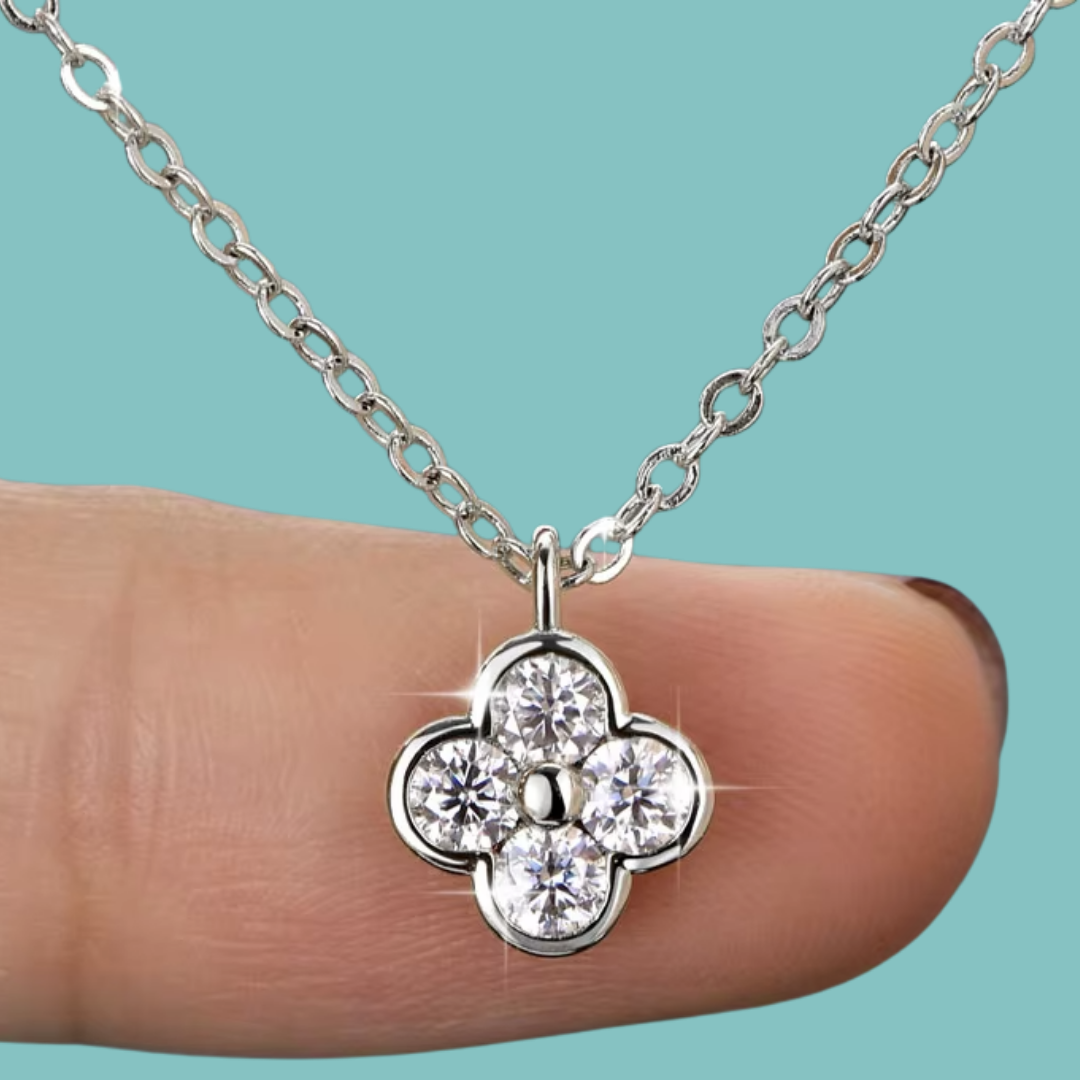 A silver-tone four-flower pendant necklace with four clear round-cut stones, shown resting on a person's fingertip against a solid turquoise background.